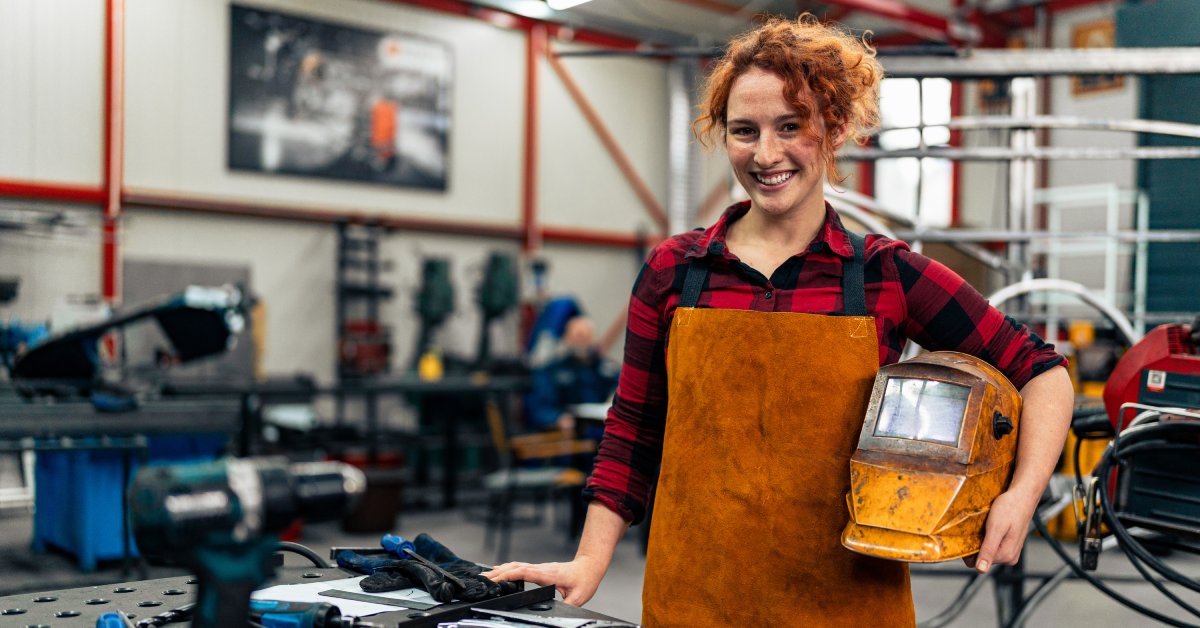 A woman is standing in a workshop and smiling at the camera. She is wearing protective equipment and holding a helmet.